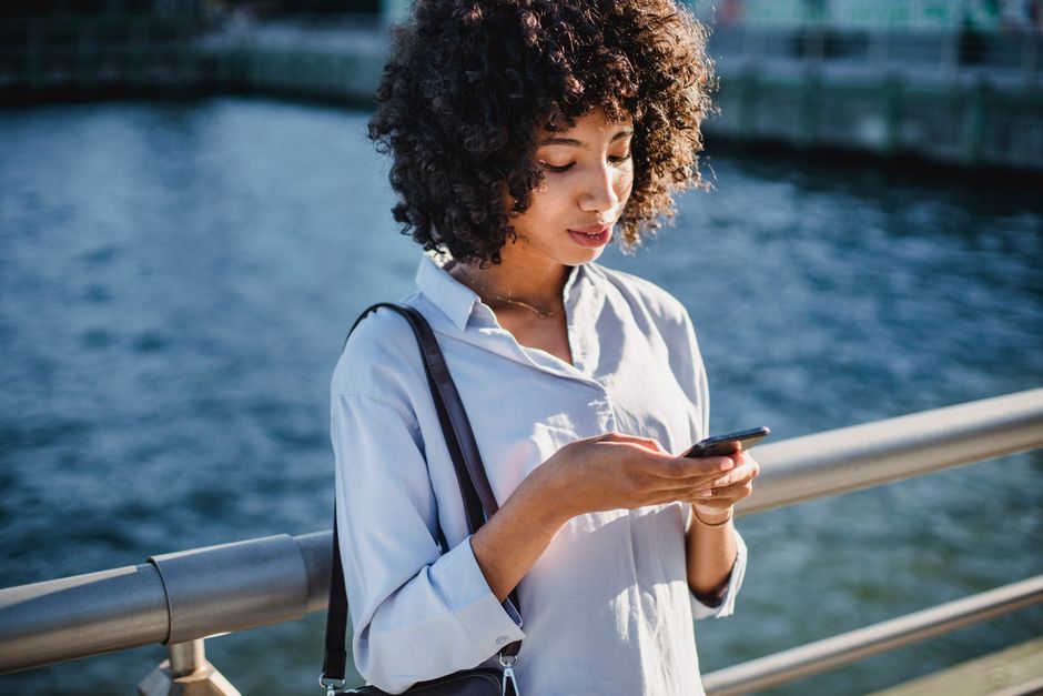 Young woman using her smartphone on a sunny day by the waterfront bridge.