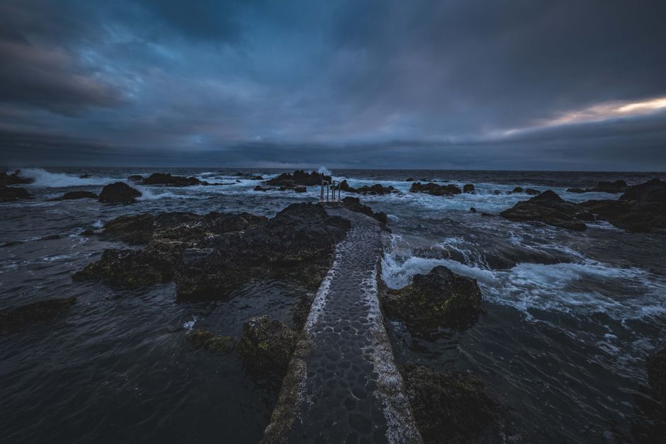 A moody seascape with a stone pathway leading into the ocean under a dramatic twilight sky.