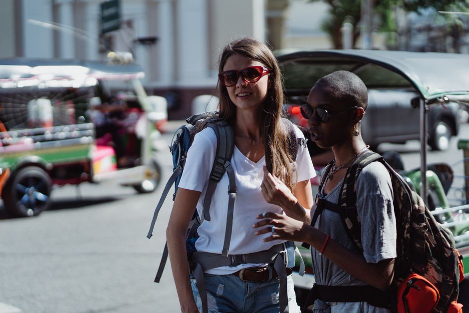 Two women travelers with backpacks and sunglasses exploring a lively street scene.