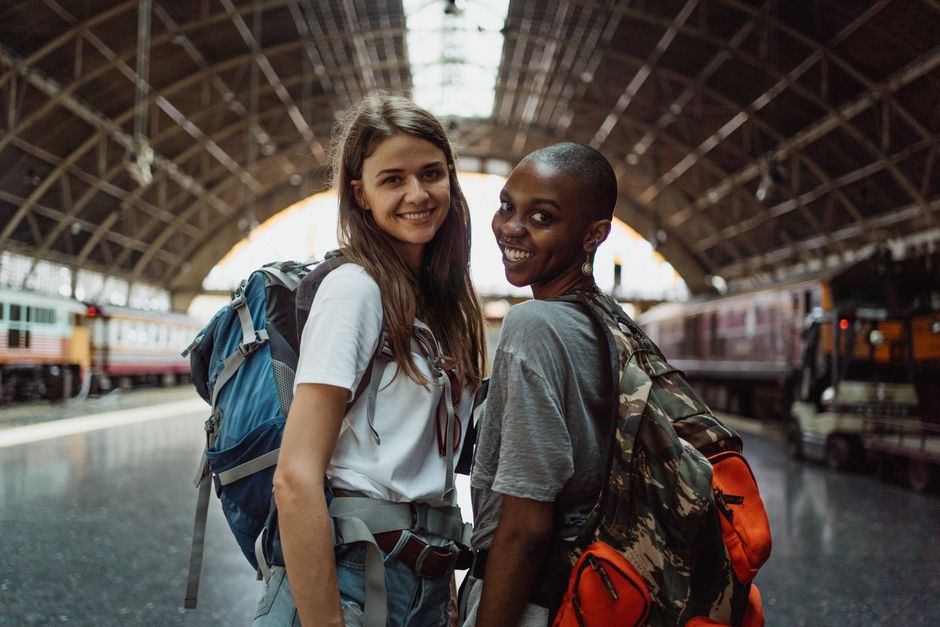 Two women with backpacks smiling at a train station, ready for their travel adventure.