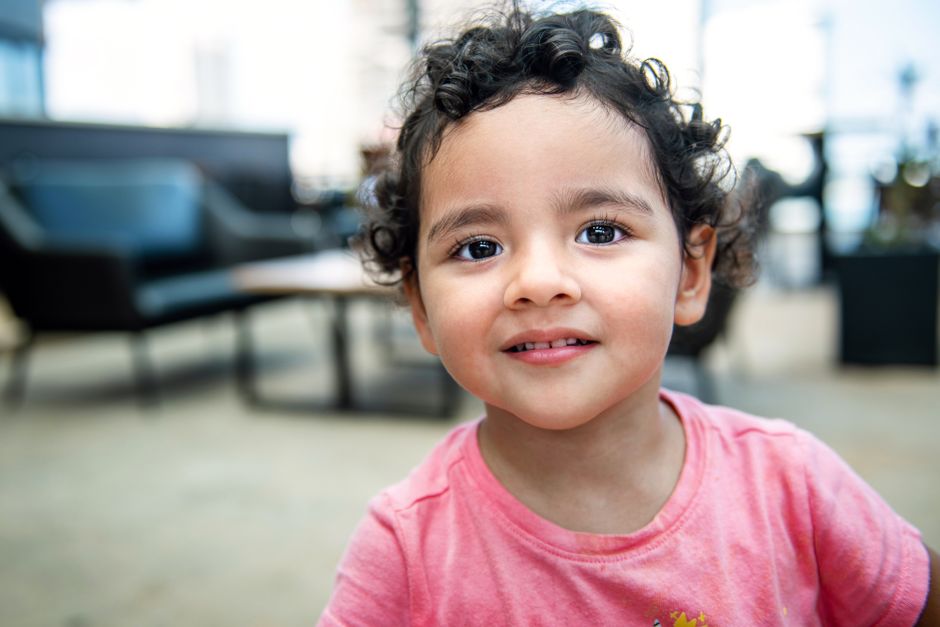 Adorable young child with curly hair smiling indoors wearing a pink shirt.