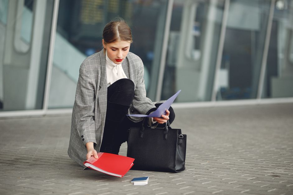 Distracted female manager in trendy wear collecting scattered documents and passport from sidewalk into briefcase near glass wall of modern building