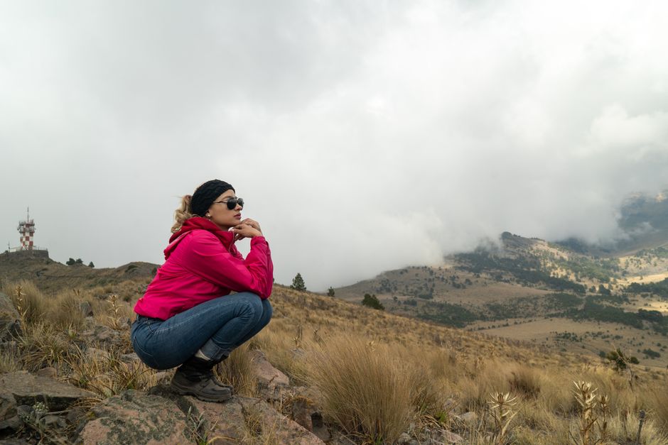 A woman in a pink jacket crouches on a rocky mountain with a cloudy view.