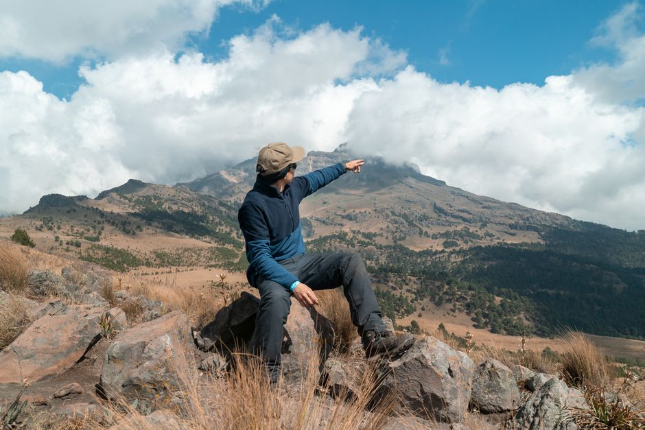Man pointing towards scenic mountainous terrain. Clear sky and rocky environment create a sense of adventure.
