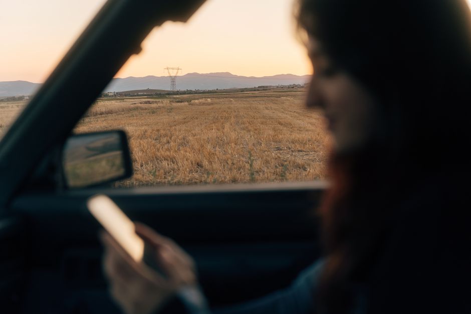 A woman inside a car uses her phone, capturing the serene sunset view outdoors.