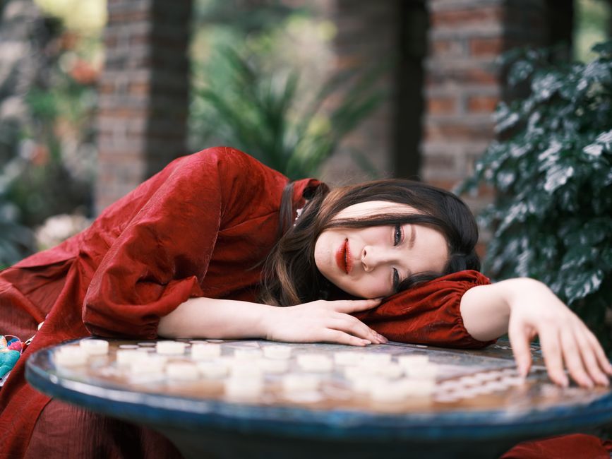 Woman in red dress resting on a table in a lush outdoor setting, serene mood.