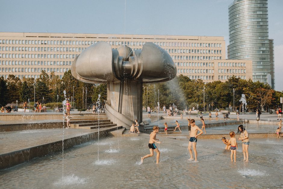 Kids enjoying a fountain in a city square with a sculpture and skyscraper in the background.