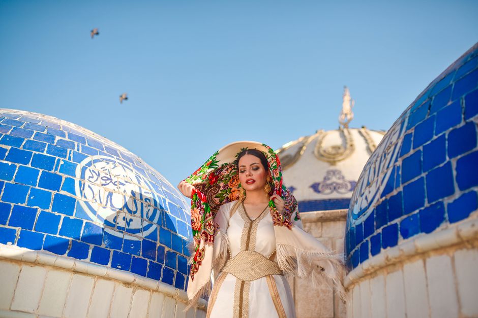 A woman in traditional clothing stands between ornate blue domes under a clear sky.