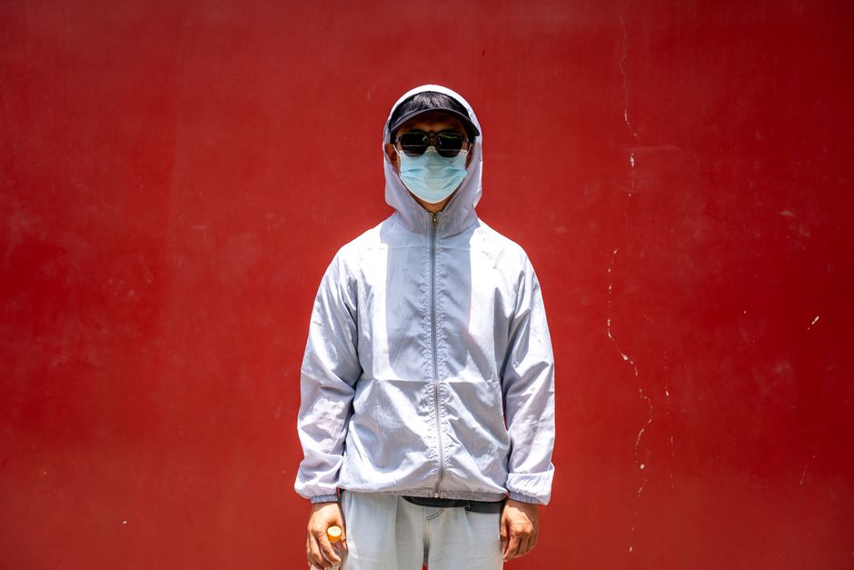 An adult wearing a mask, sunglasses, and hoodie stands against a vibrant red wall.