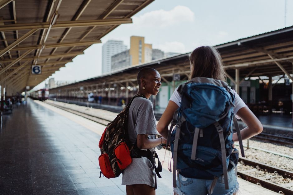 Two diverse female backpackers wait on a sunny railway platform, ready for adventure.