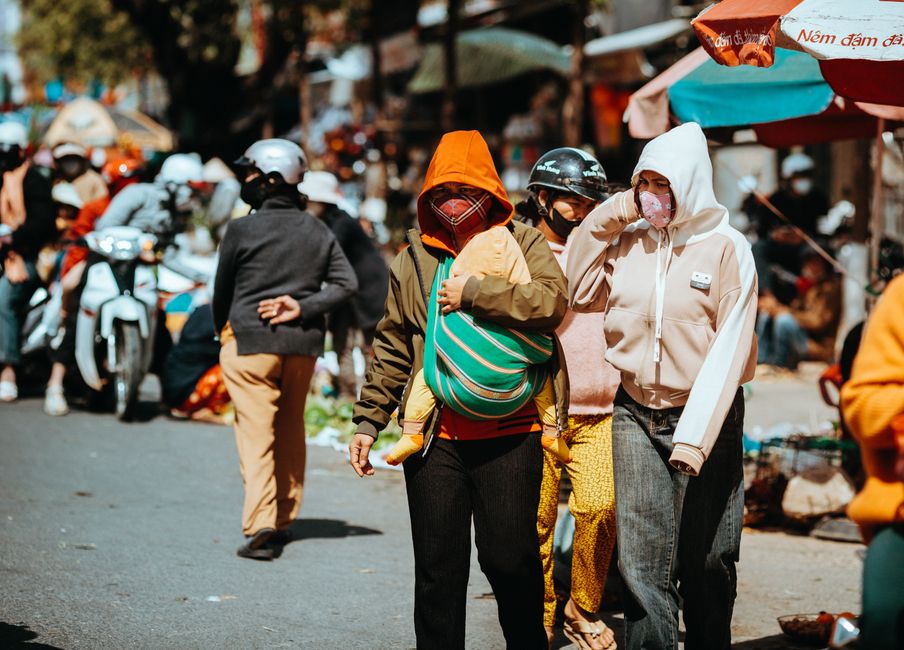 People walk through a bustling street market in Kon Tum, Vietnam, wearing face masks and colorful clothing.