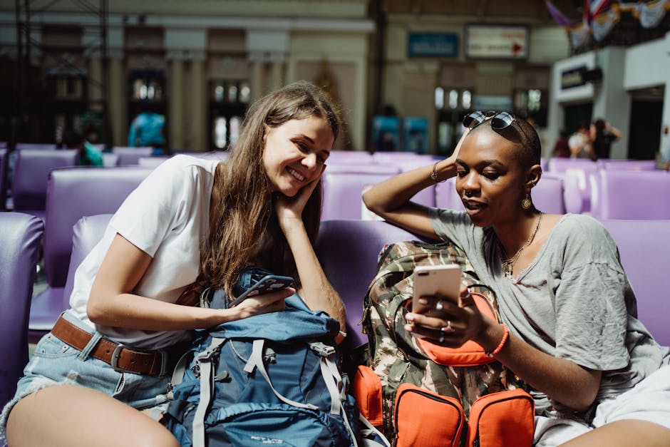 Two young women relax in an airport lounge, checking their smartphones and backpacks while waiting.