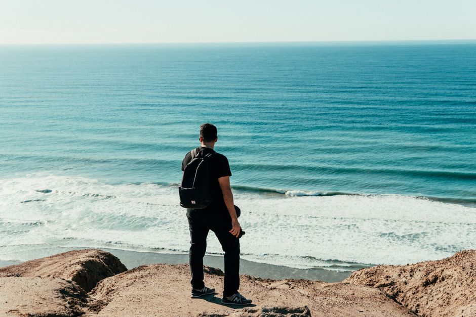A man with a backpack stands on a cliff, admiring the ocean waves below on a sunny summer day.