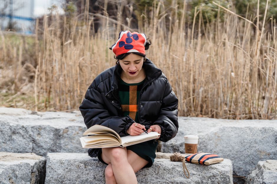 Focused young Asian female student in trendy clothes sitting with crossed legs on stone and writing notes in notebook while preparing material for project in nature in daytime