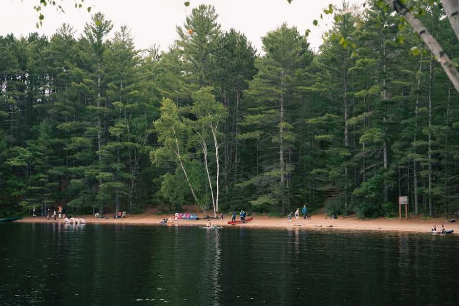 Tranquil lakeside view with pine forest, sandy beach, and people enjoying nature.