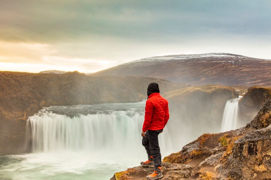 A lone traveler in a red jacket marvels at a powerful waterfall amidst a rugged landscape.